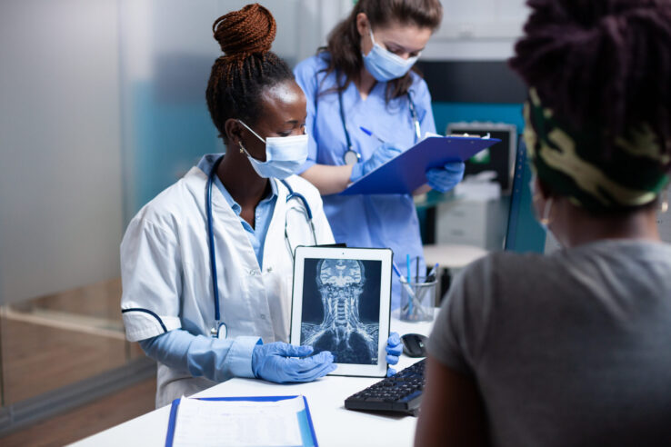 Cervical radiology specialist showing spinal ct scan results on tablet to patient with neck pain. Doctor and nurse with protective workwear gloves and face mask holding x-ray scan at medical checkup